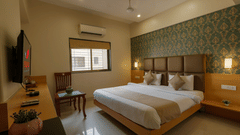 A room at Magnus Square, Viman Nagar, featuring a large double bed with beige accents, patterned green wallpaper, wall-mounted TV, wooden desk, window with blinds, and soft ambient lighting.