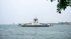 A passenger ferry in Fort Kochi, Kerala, in monsoon crosses calm coastal waters under an overcast sky, with distant ships and a faint shoreline visible on the horizon