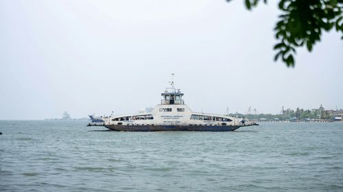 A passenger ferry in Fort Kochi, Kerala, in monsoon crosses calm coastal waters under an overcast sky, with distant ships and a faint shoreline visible on the horizon