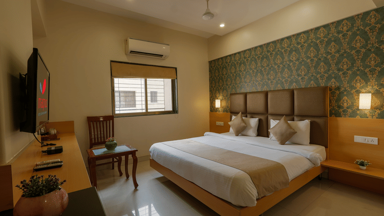 A room at Magnus Square, Viman Nagar, featuring a large double bed with beige accents, patterned green wallpaper, wall-mounted TV, wooden desk, window with blinds, and soft ambient lighting.