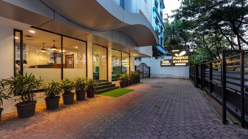 Driveway or pavement at Magnus Riverside, Kalyani Nagar featuring flower pots on one side. The ground floor of the building is made of glass walls through which we can see the interiors.
