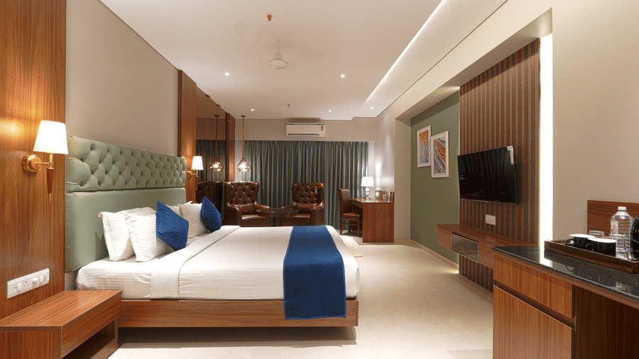 A room with a king size bed with blue accents, a wall-mounted television, wooden cabinetry, and a small sitting area at Magnus Kapitol, Sakore Nagar, Viman Nagar.