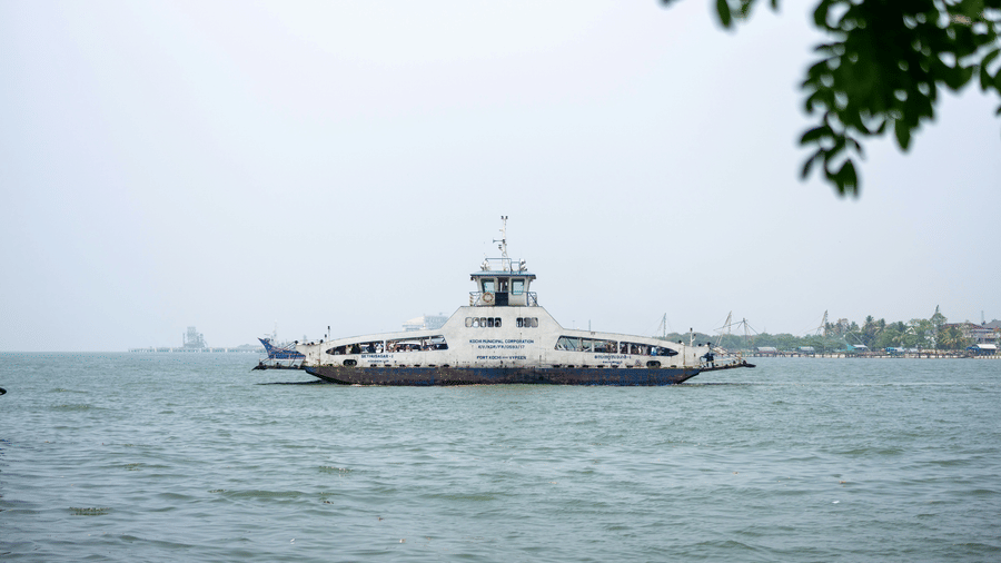 A passenger ferry in Fort Kochi, Kerala, in monsoon crosses calm coastal waters under an overcast sky, with distant ships and a faint shoreline visible on the horizon