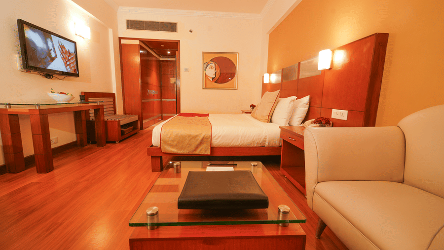 Luxury suite at The Suryaa New Delhi featuring a large bed, sofa, glass coffee table, work desk, and a wall-mounted television