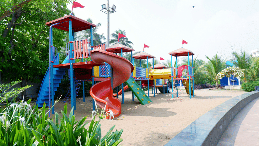 children’s playground with slides, climbing frames, bridges and towers set on sand, surrounded by trees and garden landscaping