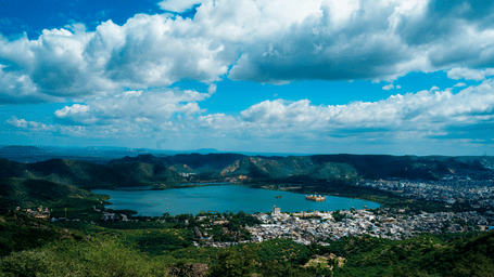 Scenic view of Jal Mahal surrounded by lush green hills beneath bright monsoon skies, with Jaipur spread across the shoreline.
