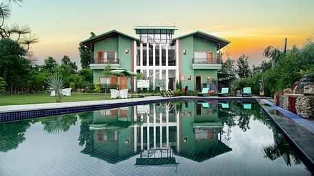 A modern resort building reflected in a large swimming pool, with a vibrant sunset sky in the background.