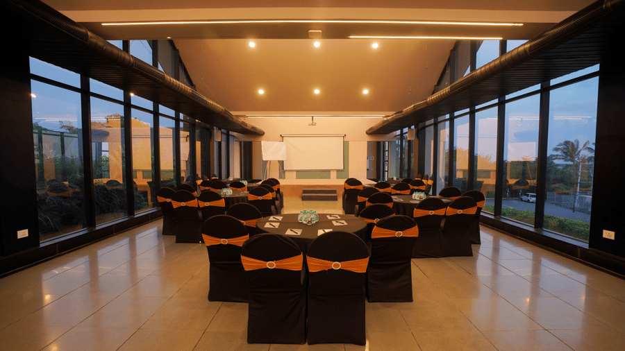 Blue Moon event hall at Darza Resorts arranged for a conference with round tables, projector screen, stage and black and orange seating overlooking the outdoors in Coimbatore.