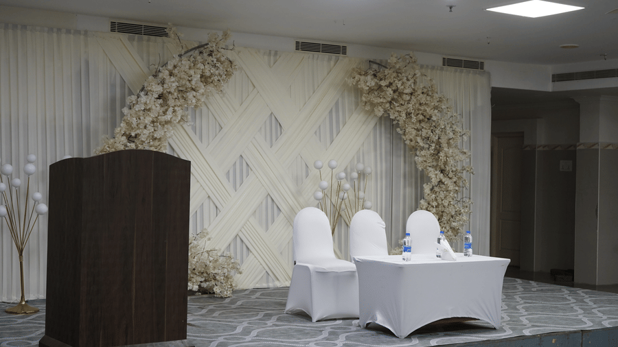 A decorated event stage featuring a wooden podium, floral backdrop decoration, and a covered table with chairs at DSF Grand Plaza, Tuticorin.