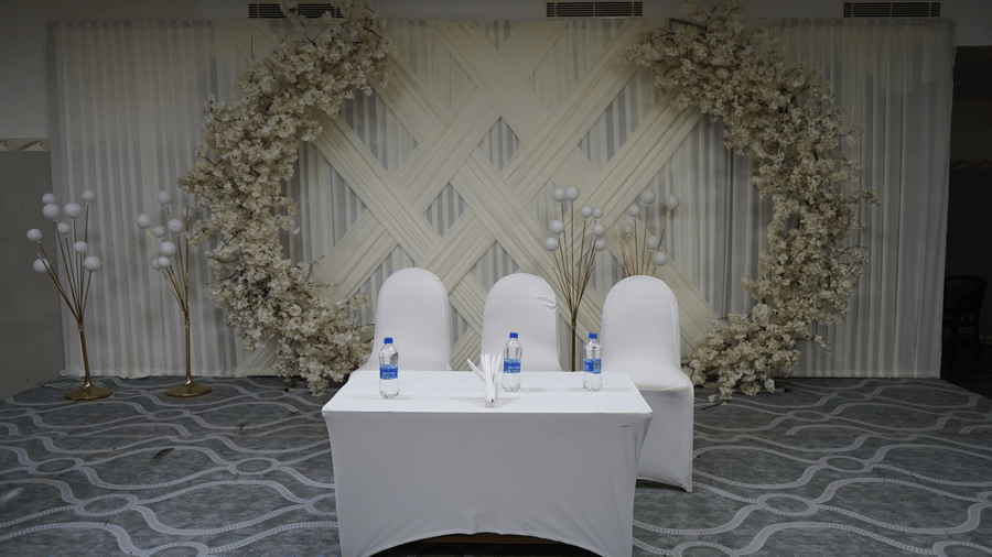 A front view of a stage setup featuring a floral arch backdrop, covered table with water bottles, and chairs at DSF Grand Plaza, Tuticorin.