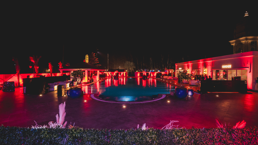 Night-time pool party with vibrant red and blue lighting illuminating the water and a nearby bar area at Hotel Hukam's Lalit Mahal.