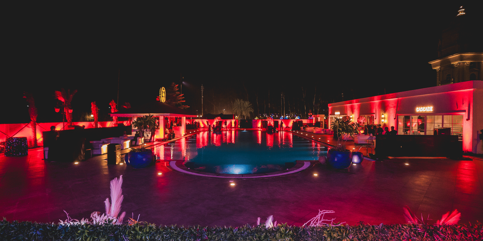 Night-time pool party with vibrant red and blue lighting illuminating the water and a nearby bar area at Hotel Hukam's Lalit Mahal.