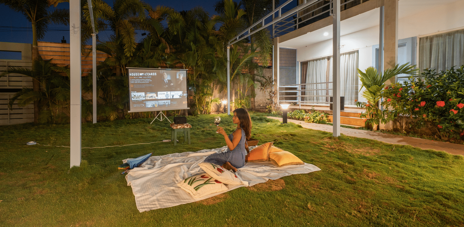 A woman enjoying an open-air movie under the stars, holding a wine glass while sitting on a picnic blanket surrounded by pillows - Perfectstayz Koko Maya