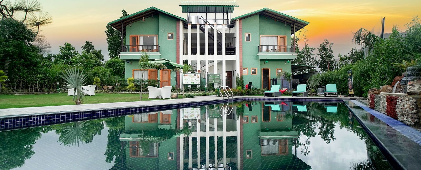 A modern resort building reflected in a large swimming pool, with a vibrant sunset sky in the background.