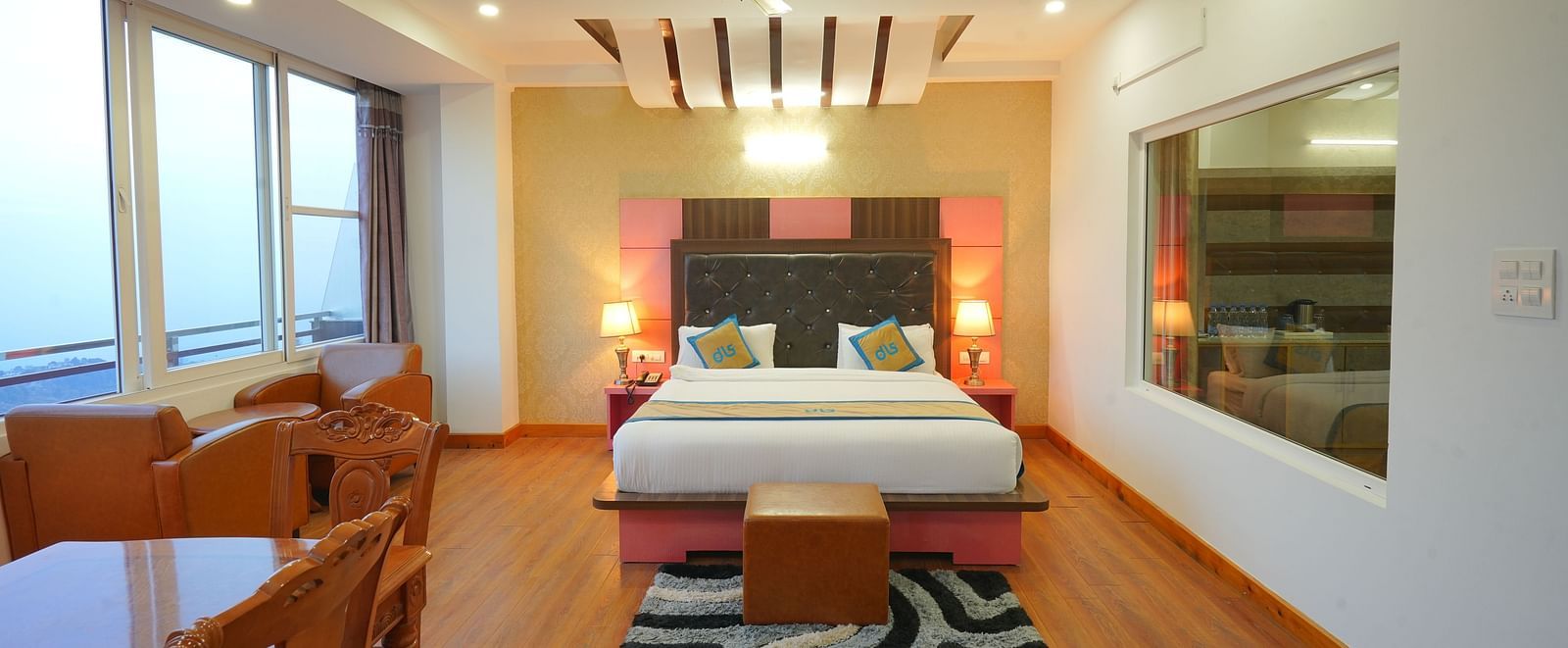 A spacious hotel room with a large bed, a seating area with a table and chairs, and large windows offering a view of the outdoors | DLS Hotel Shiva Sanctuary & Spa, Dharamshala