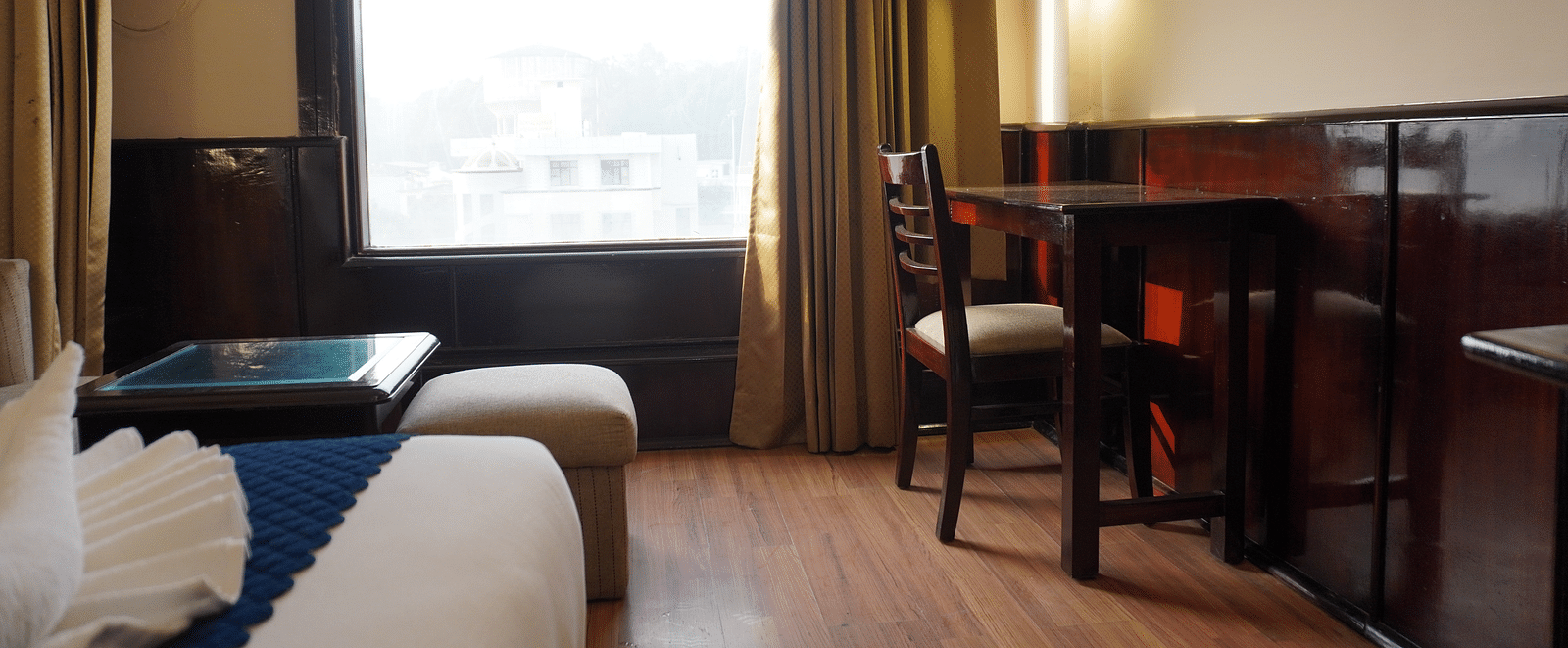 A bedroom with a chair and a table, bathed in light streaming through the window at DLS Park Grace, Haridwar.