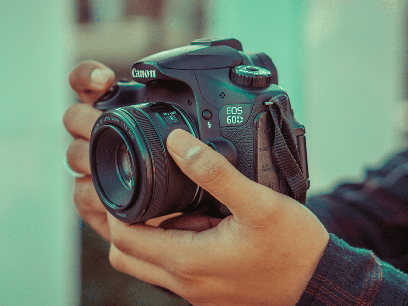 A pair of hands holds a DSLR camera with the lens facing forward against a softly blurred background.