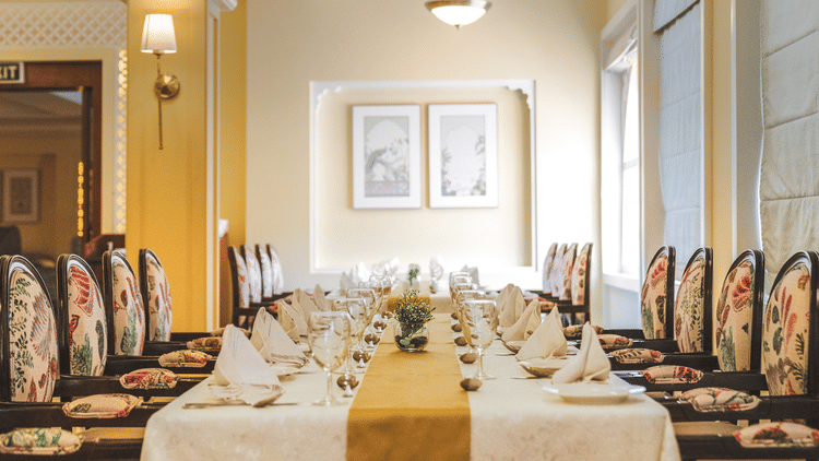 Suryamahal at The Garden Ananta Elite showcases an opulent dining space adorned with white flower symbols on the walls, an abundance of chandeliers, and inviting cushioned wooden chairs arranged around an elegant dining table.