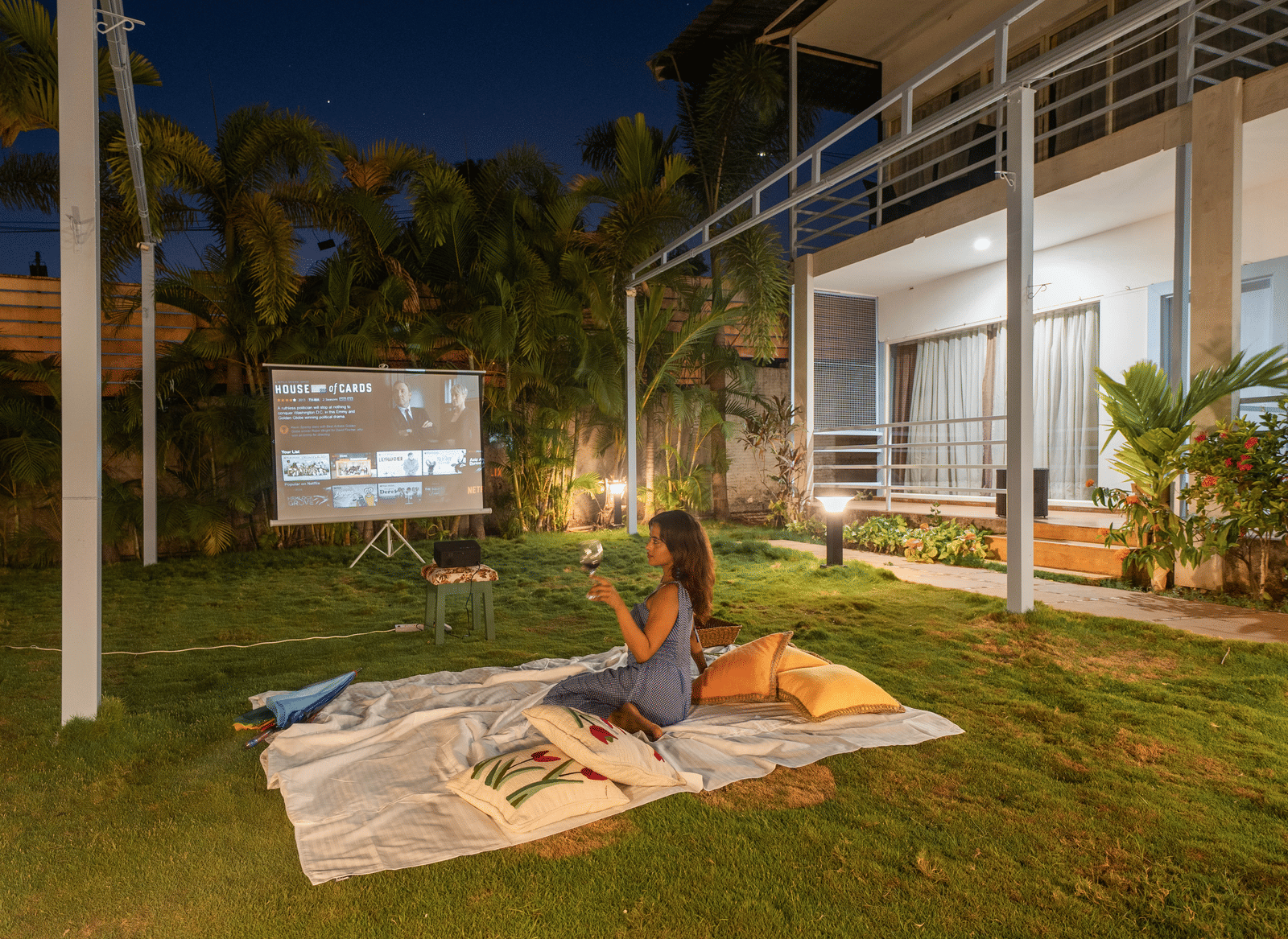 A woman enjoying an open-air movie under the stars, holding a wine glass while sitting on a picnic blanket surrounded by pillows - Perfectstayz Koko Maya