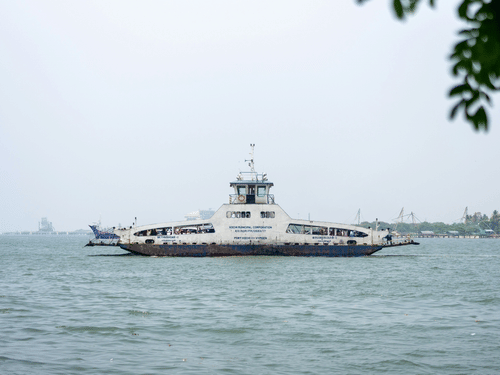 A passenger ferry in Fort Kochi, Kerala, in monsoon crosses calm coastal waters under an overcast sky, with distant ships and a faint shoreline visible on the horizon