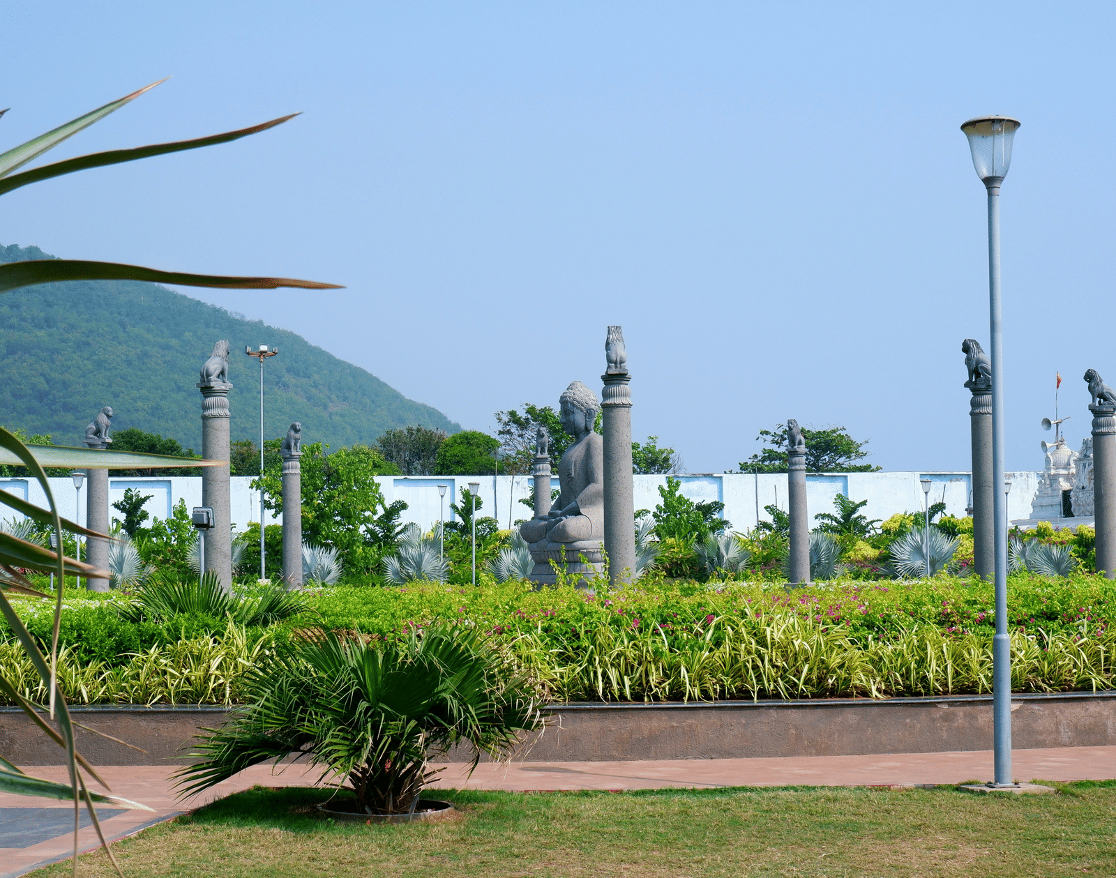 A park area with vibrant green shrubs, stone statues, a lamp post, and hills visible under a bright blue sky.