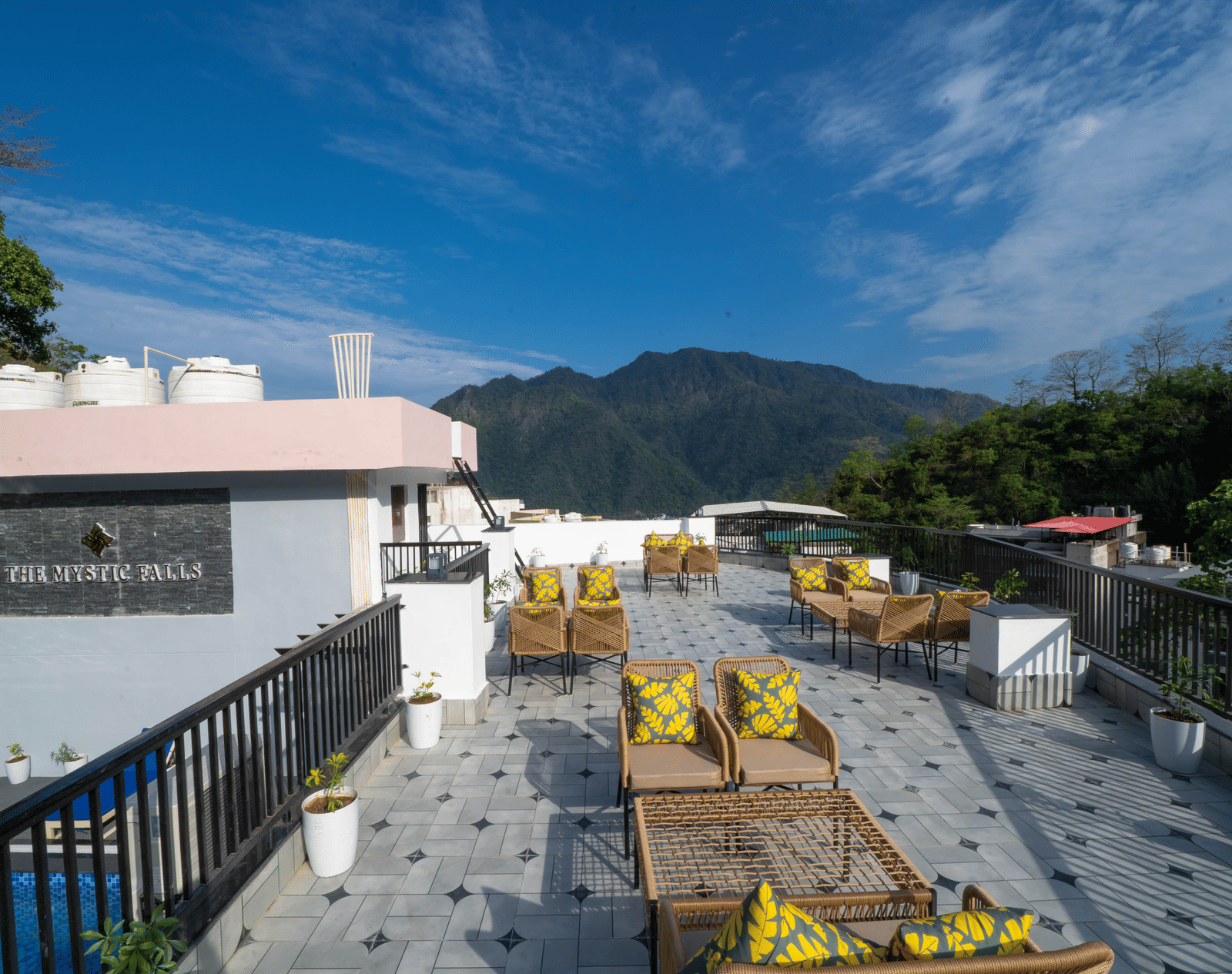 An outdoor terrace with a floor, a building structure, tables, chairs, cushions, and a view of the sky and hills in Perfectstayz Premium Mystic Falls, Rishikesh.
