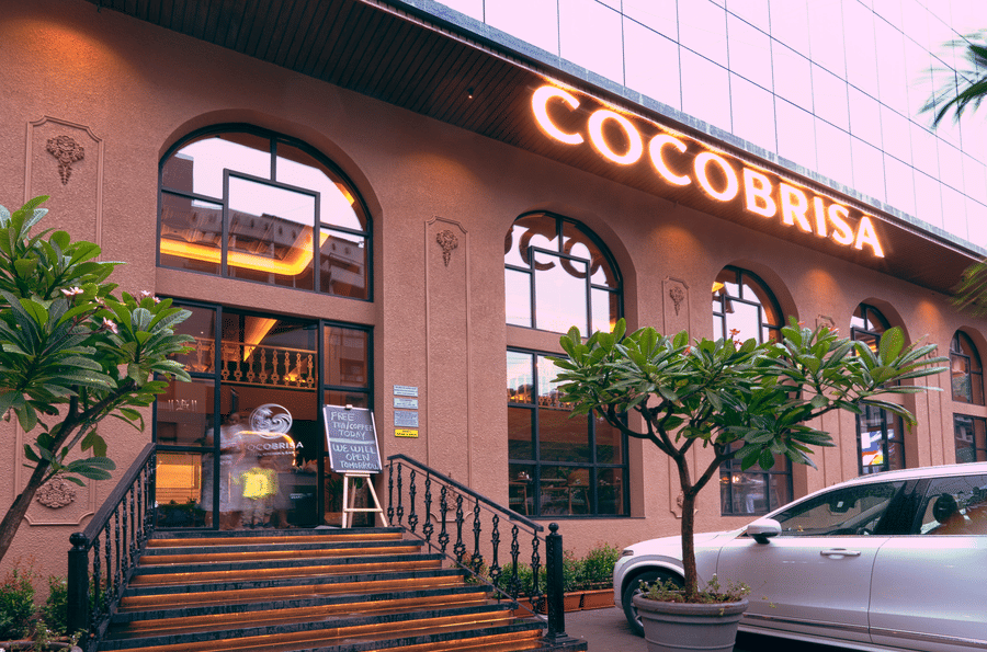 Garden dining area at Coco Brisa with contemporary tables and chairs, lush tropical plants, decorative hanging lights, fabric ceiling drapes, and a warm, inviting outdoor ambience.