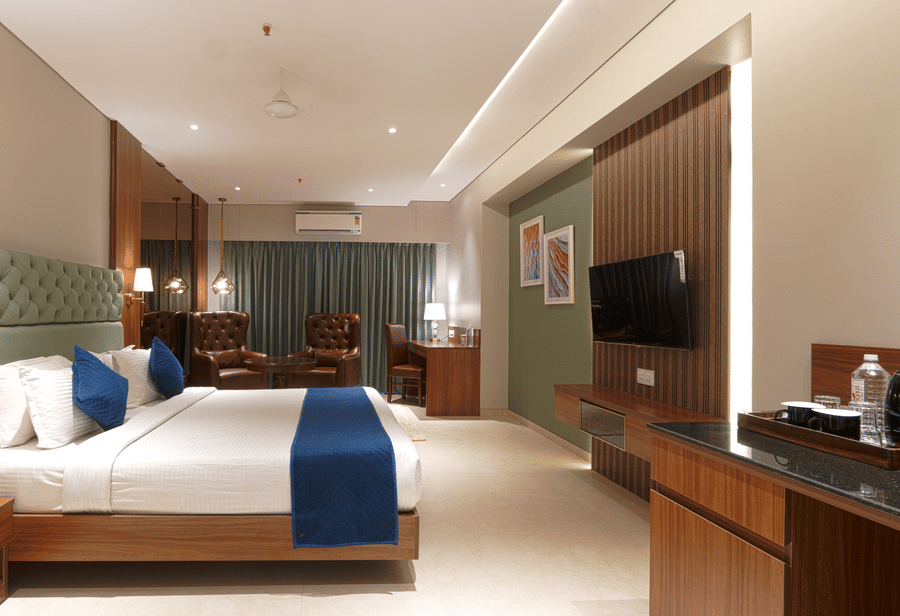 A room with a king size bed with blue accents, a wall-mounted television, wooden cabinetry, and a small sitting area at Magnus Kapitol, Sakore Nagar, Viman Nagar.