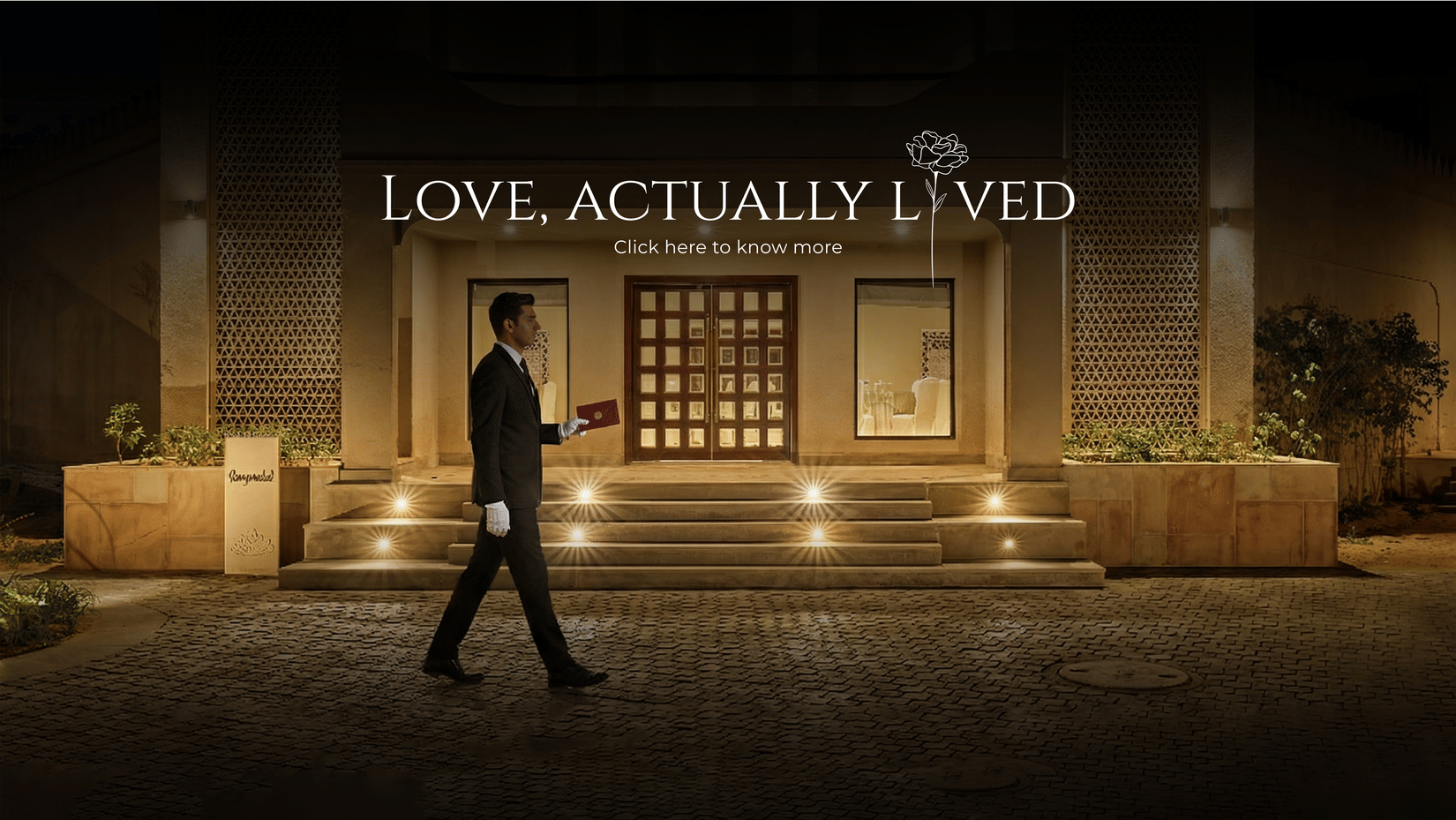 Elegant hotel entrance at night with warm lighting and a staff member walking past, showcasing the “Love, Actually Lived” romantic experience.