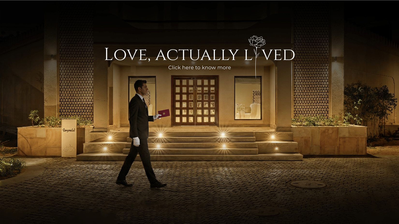 Elegant hotel entrance at night with warm lighting and a staff member walking past, showcasing the “Love, Actually Lived” romantic experience.