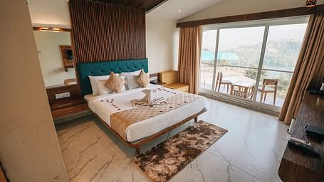 An image of a room with a bed, bed side tables, an expansive glass door leading to the balcony where a table and chairs are placed - Nisarga Lake Resort