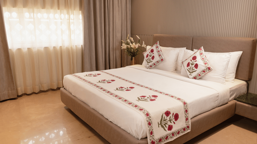 A spacious bedroom with a king size bed dressed in embroidered linen, decorative cushions, a broad upholstered headboard, bedside tables on either side, soft window drapes, and a floral arrangement placed near the bed.