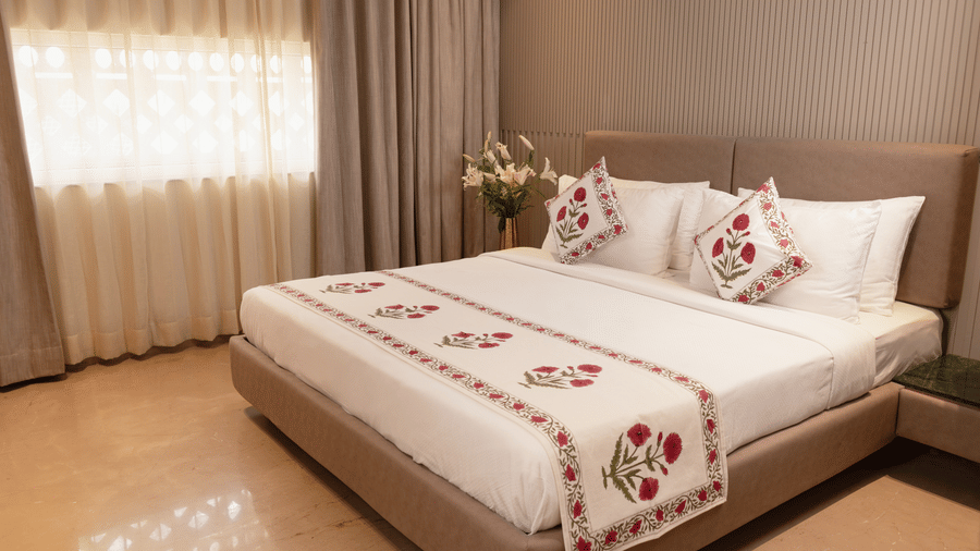 A spacious bedroom with a king size bed dressed in embroidered linen, decorative cushions, a broad upholstered headboard, bedside tables on either side, soft window drapes, and a floral arrangement placed near the bed.