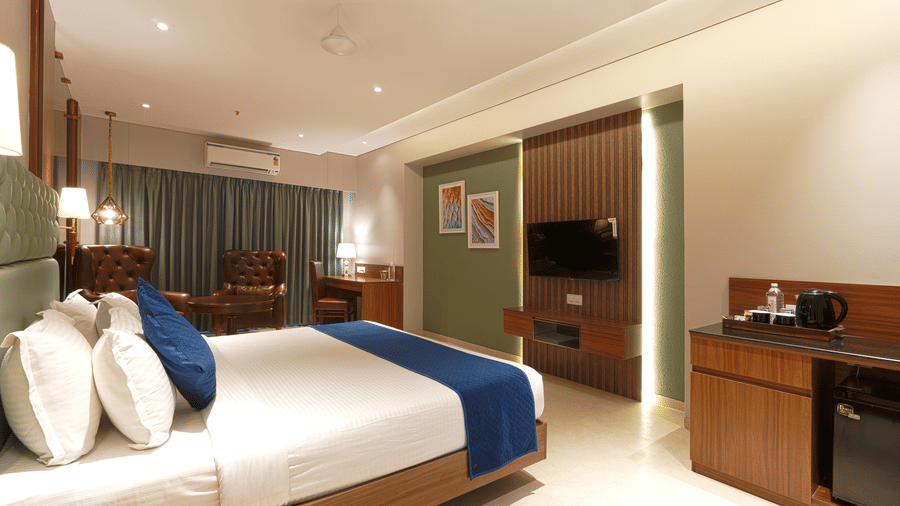 A room with a king size bed showing white pillows, a blue bed runner, a television, and a wooden desk at Magnus Kapitol, Sakore Nagar, Viman Nagar.