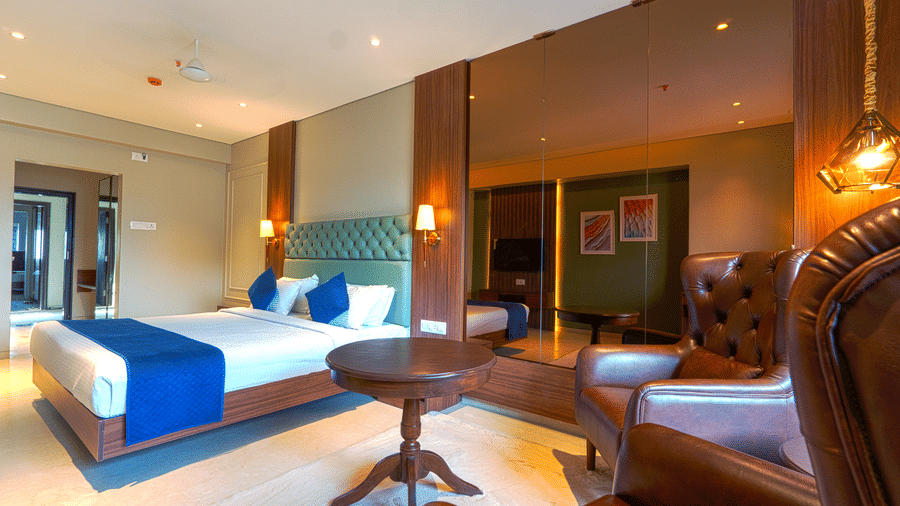 A room with a bed featuring blue pillows, a wooden side table, and a leather seating area in the background at Magnus Kapitol, Sakore Nagar, Viman Nagar.