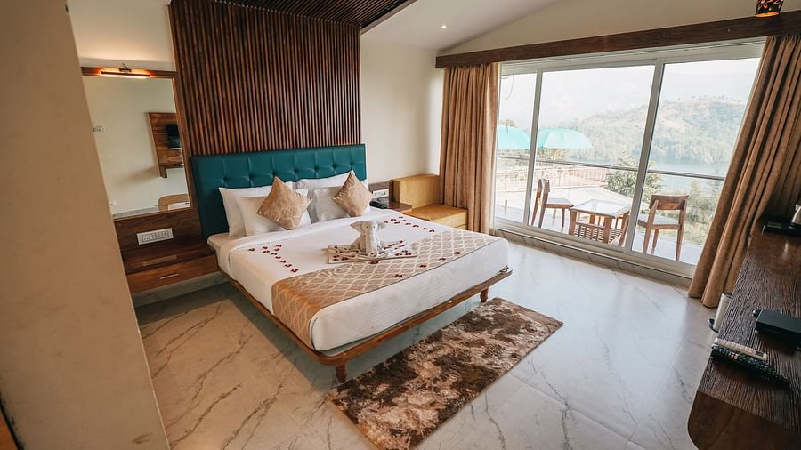 An image of a room with a bed, bed side tables, an expansive glass door leading to the balcony where a table and chairs are placed - Nisarga Lake Resort