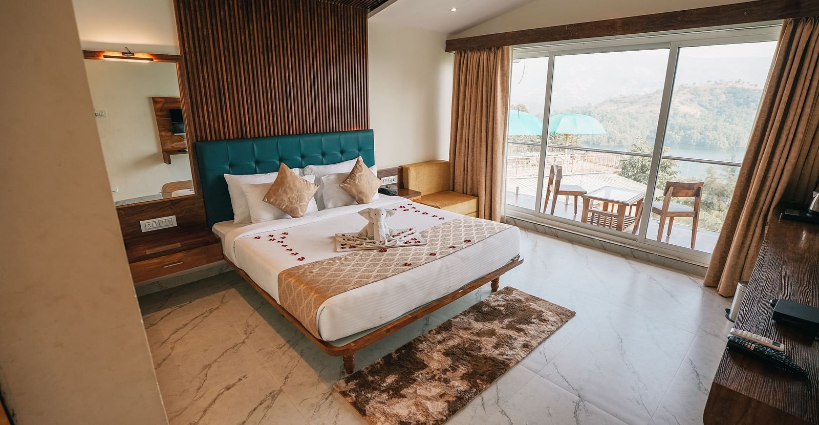 An image of a room with a bed, bed side tables, an expansive glass door leading to the balcony where a table and chairs are placed - Nisarga Lake Resort