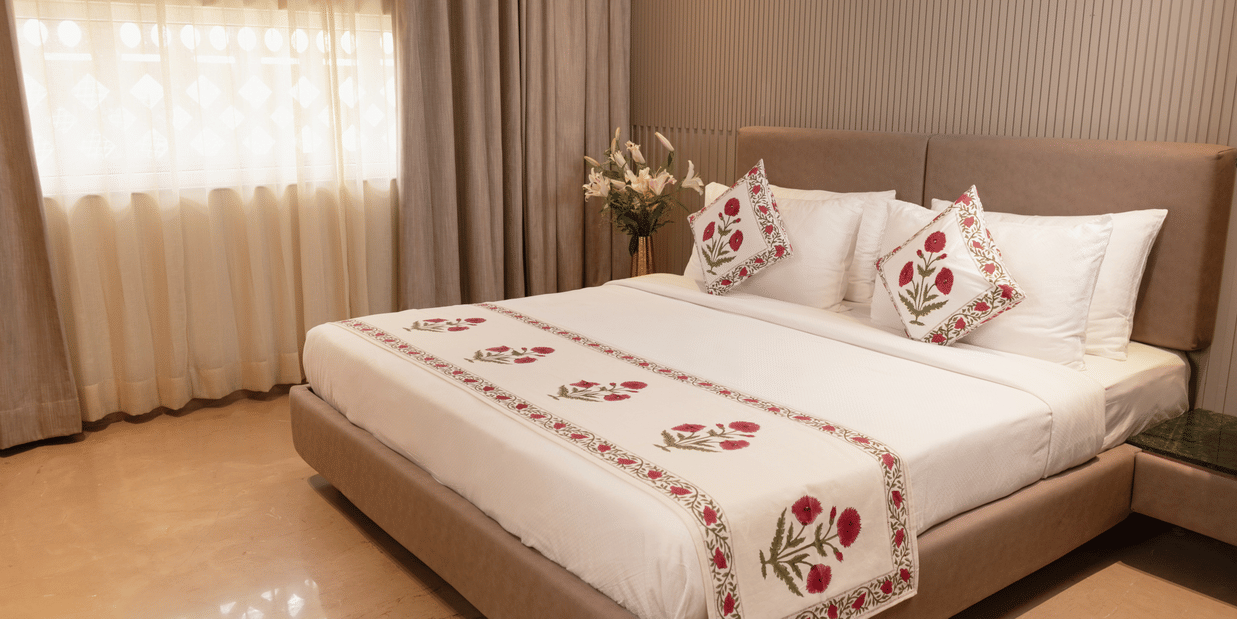 A spacious bedroom with a king size bed dressed in embroidered linen, decorative cushions, a broad upholstered headboard, bedside tables on either side, soft window drapes, and a floral arrangement placed near the bed.