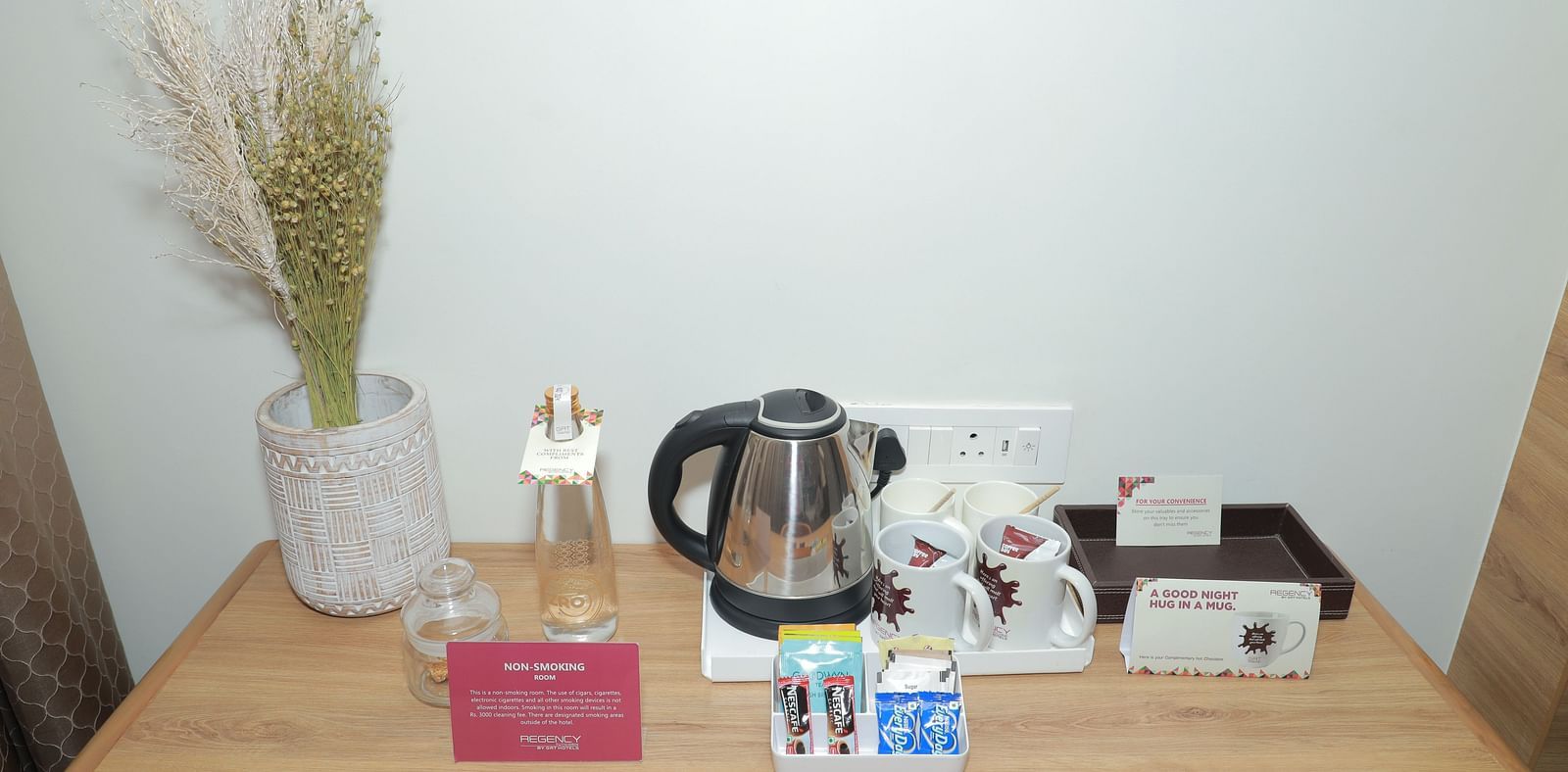An electric kettle, instant coffee and tea sachets, a water bottle, and a vase placed on a desk in our Regent Suites in Tuticorin - Regency Tuticorin by GRT Hotels
