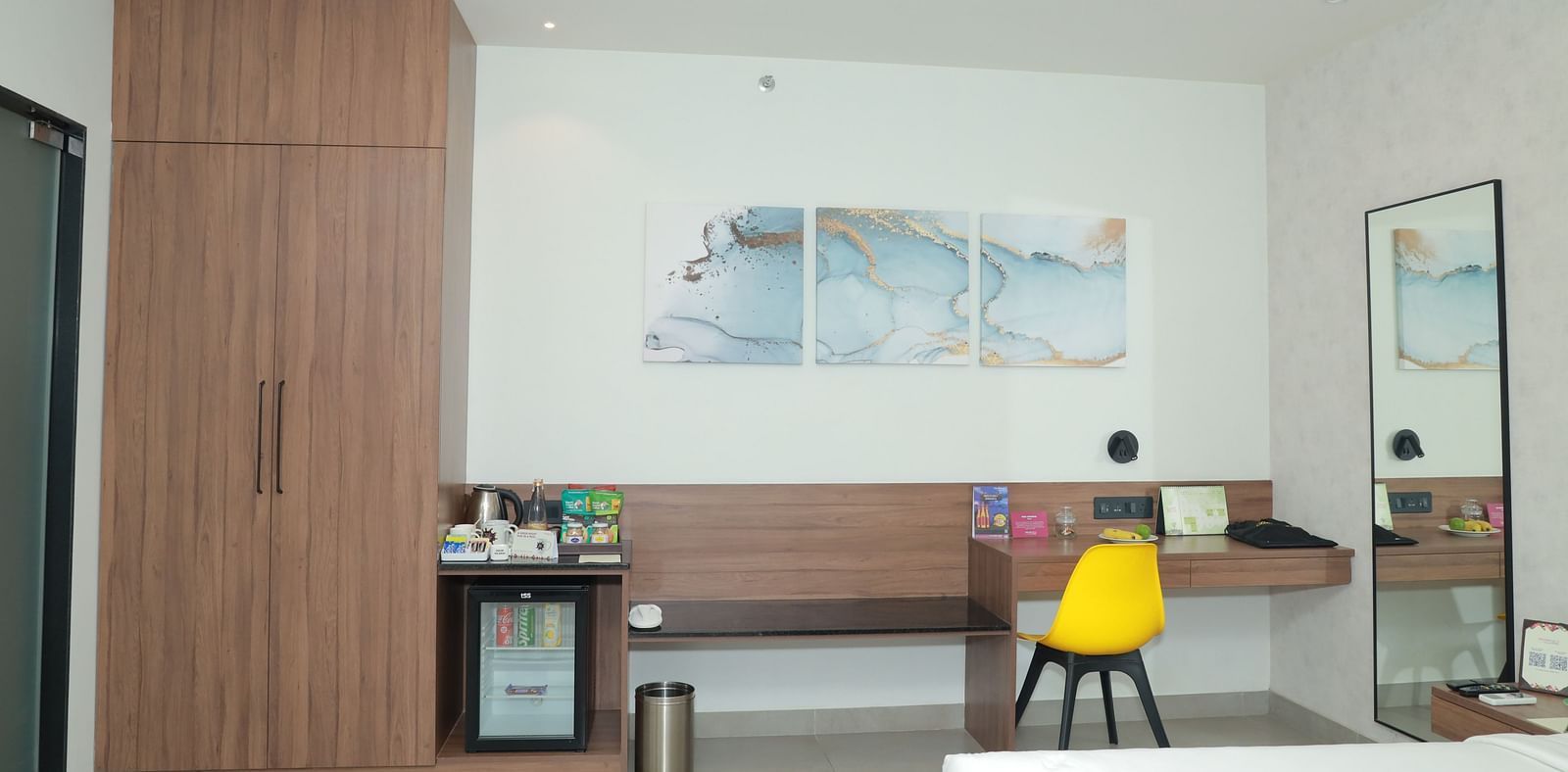 An image of a work area in our suite in Tuticorin, with a yellow chair, a desk with snacks, a wooden cupboard, and a mirror - Regency Tuticorin by GRT Hotels