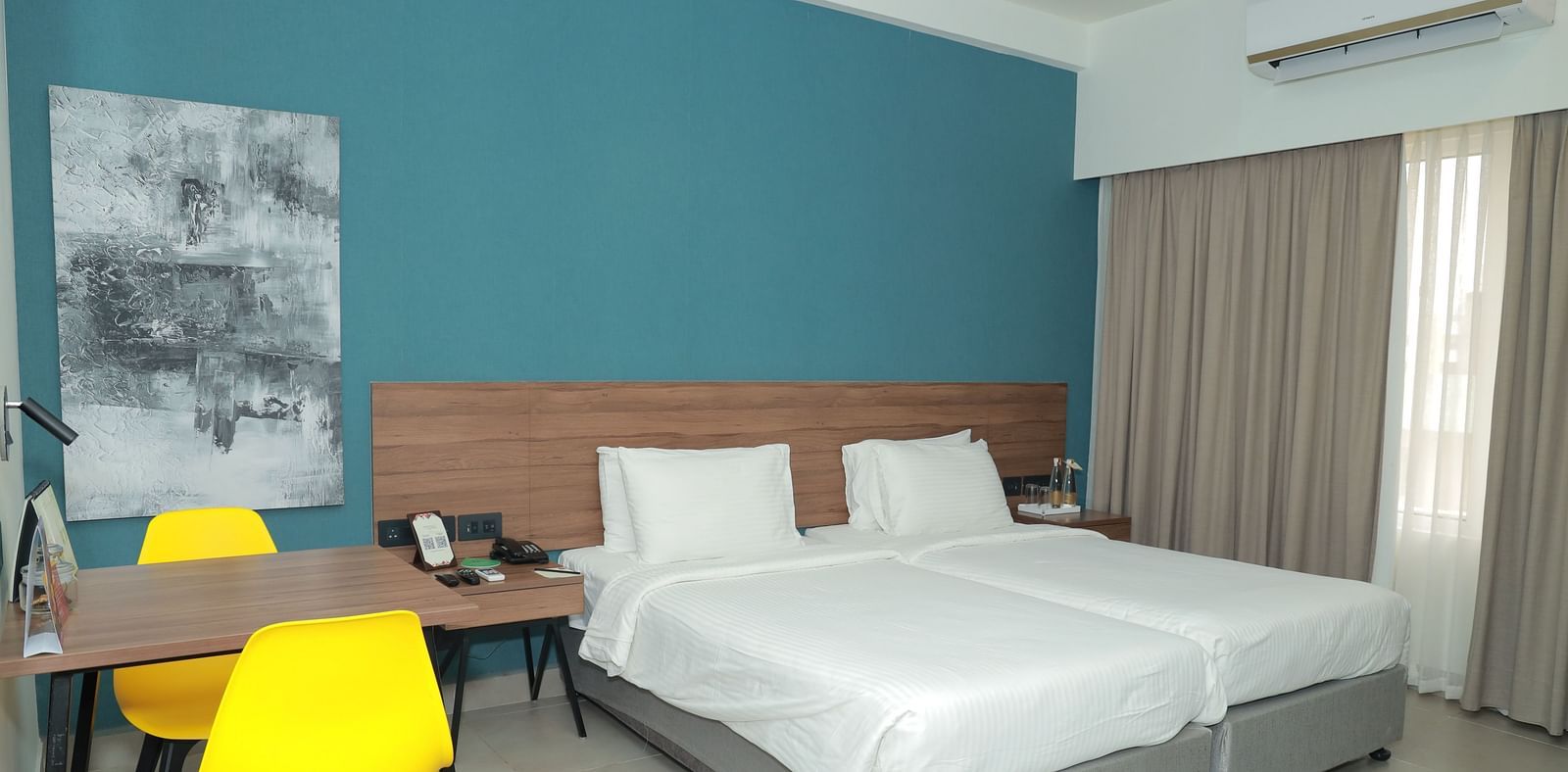 An image of our Deluxe Rooms featuring twin beds, a table with two yellow chairs, and a painting hanging on the wall beside the bed - Regency Tuticorin by GRT Hotels
