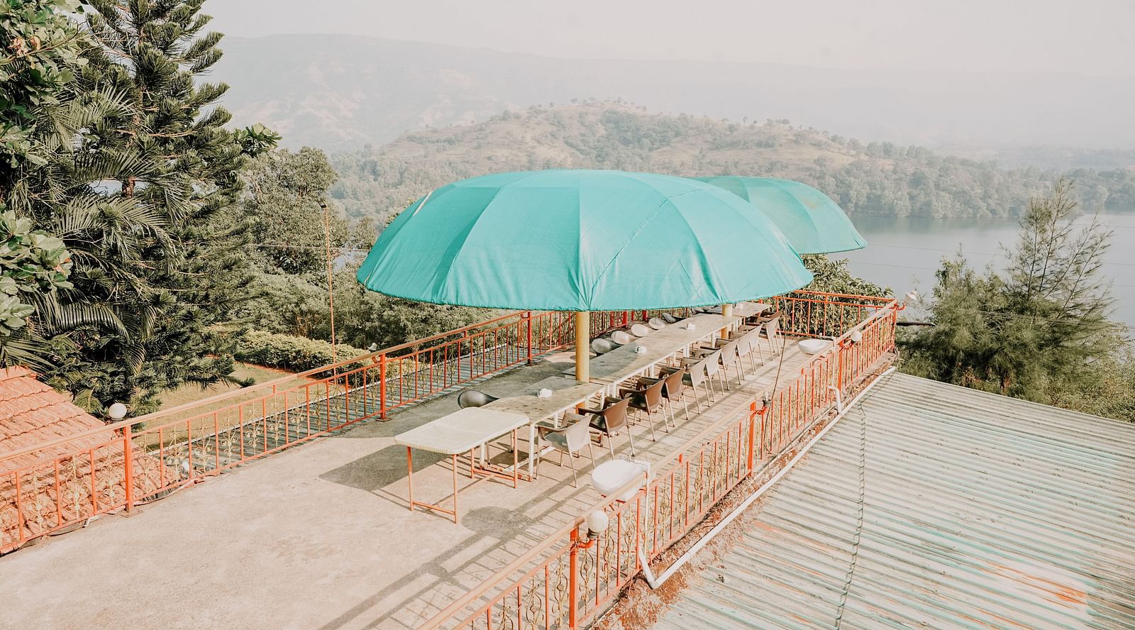 An image of a rooftop bar at Nisarga Lake Resort featuring chairs, tables and umbrellas, offering views of the mountains and lake