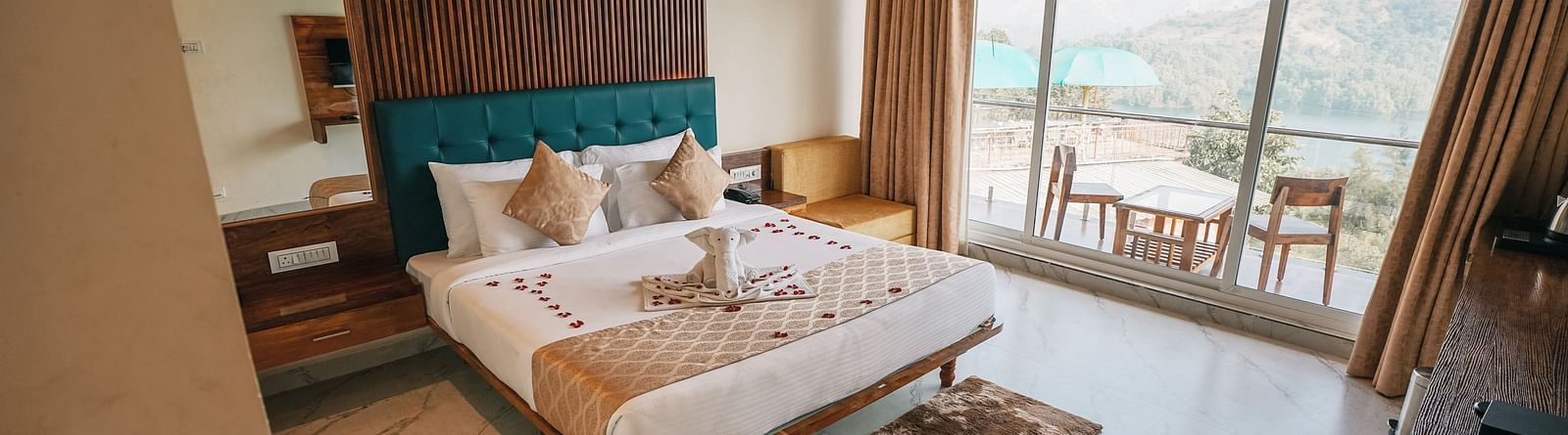 An image of a room with a bed, bed side tables, an expansive glass door leading to the balcony where a table and chairs are placed - Nisarga Lake Resort