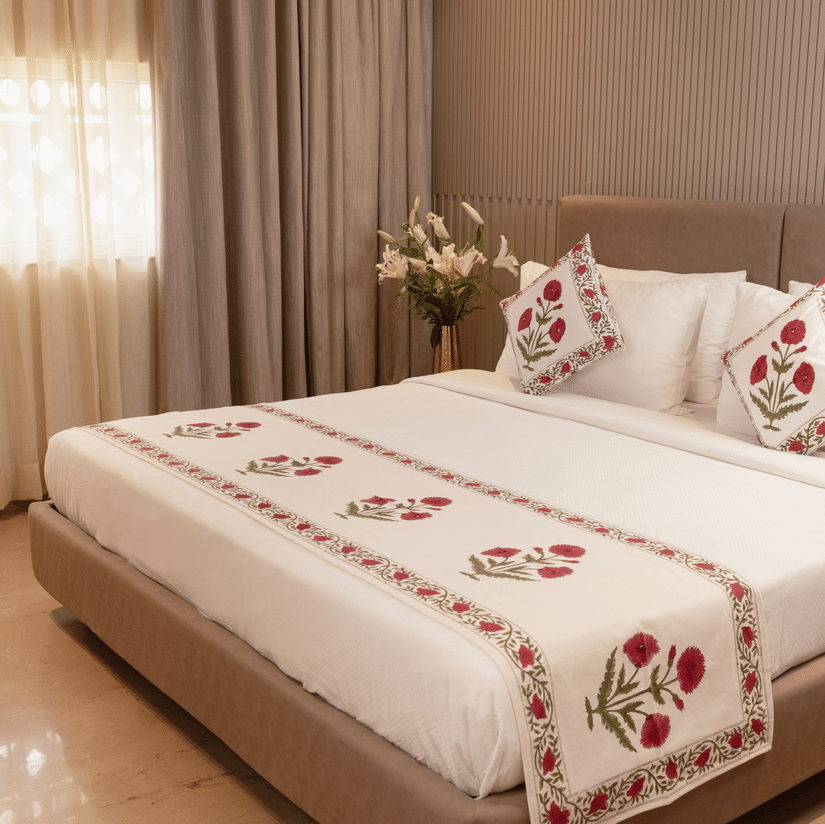 A spacious bedroom with a king size bed dressed in embroidered linen, decorative cushions, a broad upholstered headboard, bedside tables on either side, soft window drapes, and a floral arrangement placed near the bed.