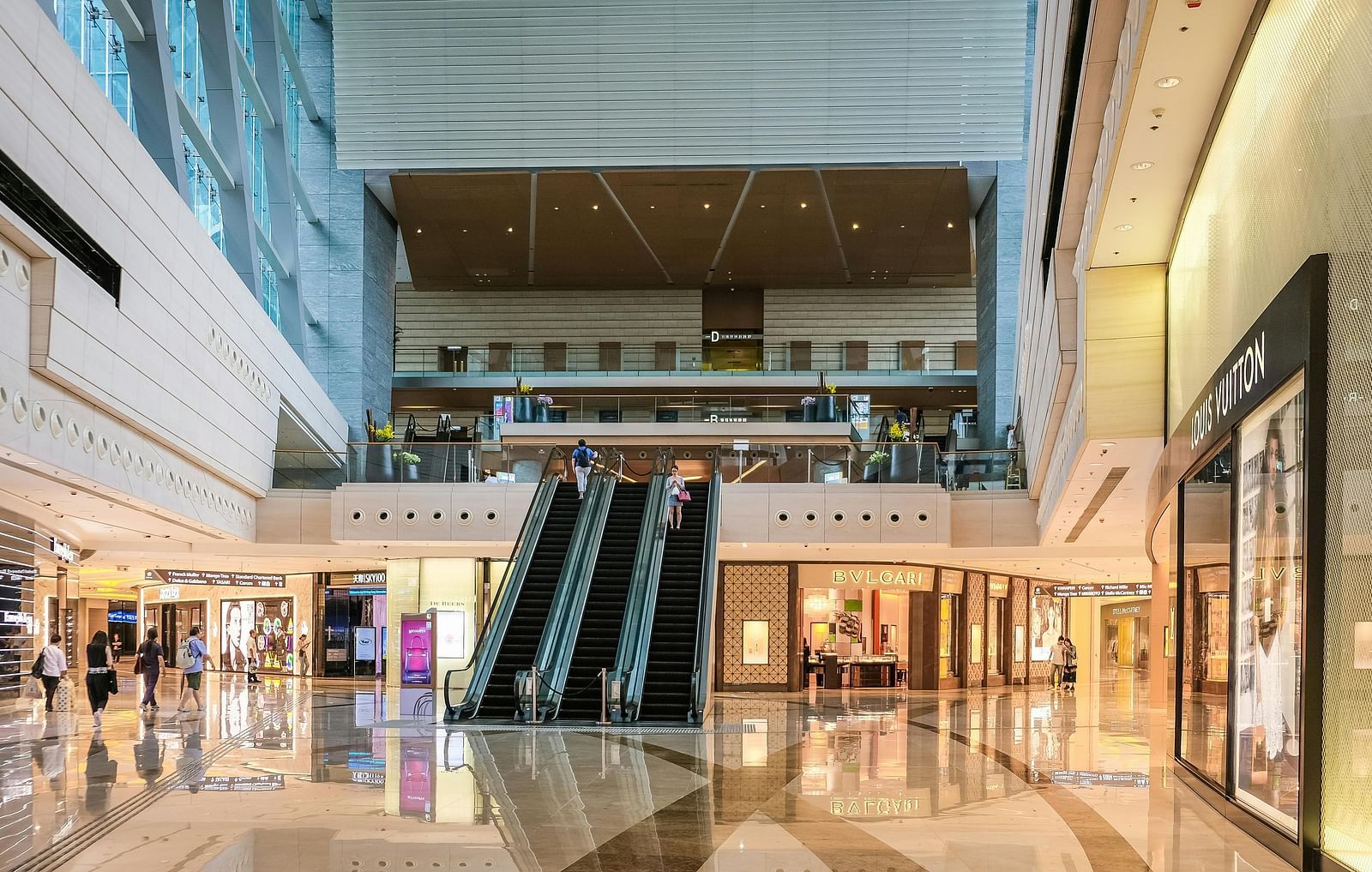 Interior view of a shopping mall with two escalators in the center, leading to an upper level. Shops line the polished floor on either side.