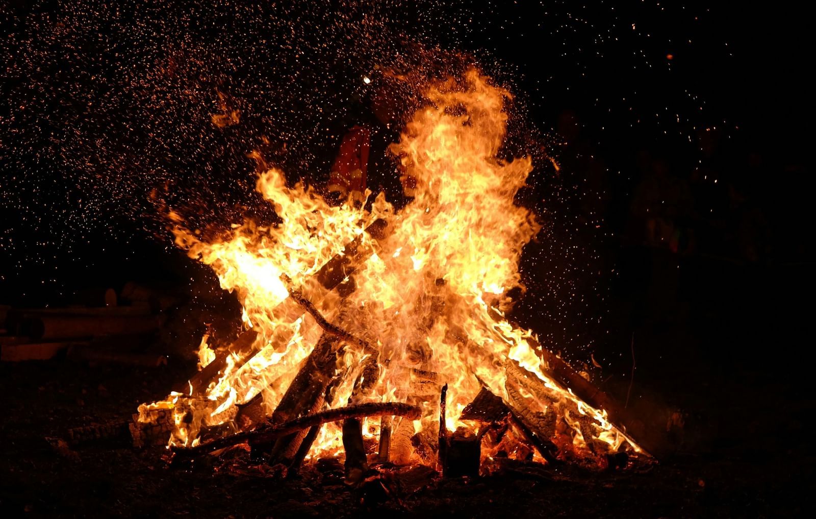 A bright bonfire with orange and red flames burns brightly in the dark. Sparks fly from the burning logs.