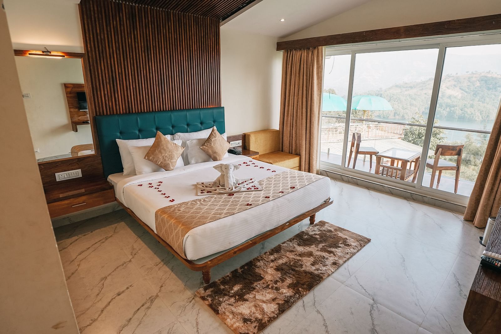 An image of a room with a bed, bed side tables, an expansive glass door leading to the balcony where a table and chairs are placed - Nisarga Lake Resort
