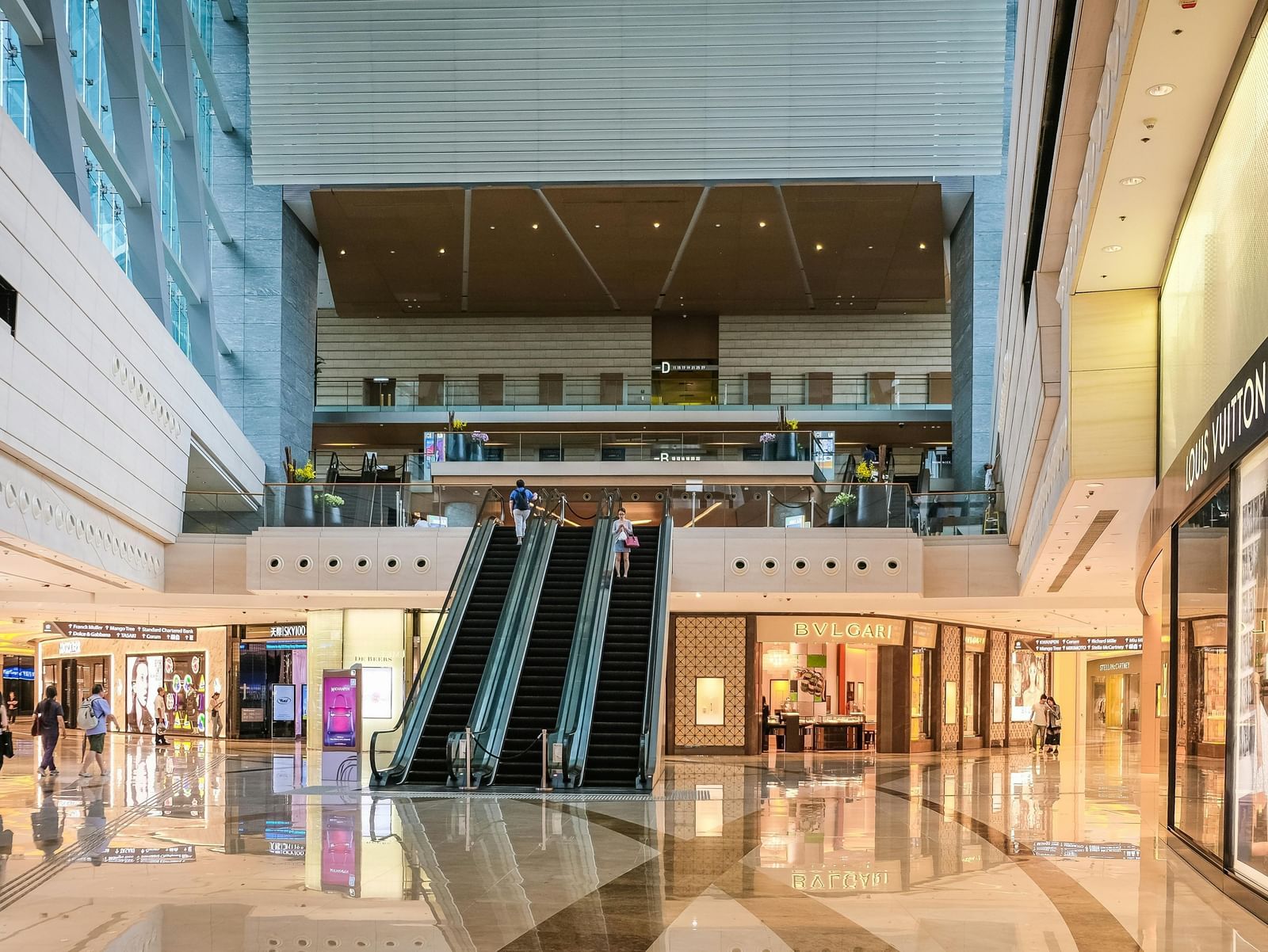 Spacious shopping mall interior with escalators, glass railings and bright store displays.