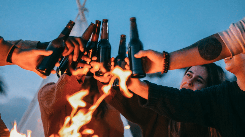 Several people raise their beer bottles in a toast around an outdoor fire at night.