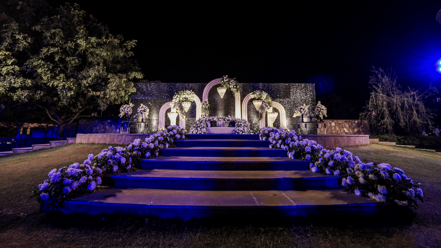 At one of the best Banquet Halls in Jaipur, a wedding stage is set at Ananta Spa & Resort, Jaipur, is viewed across blue-lit steps adorned with purple and white flowers at night.